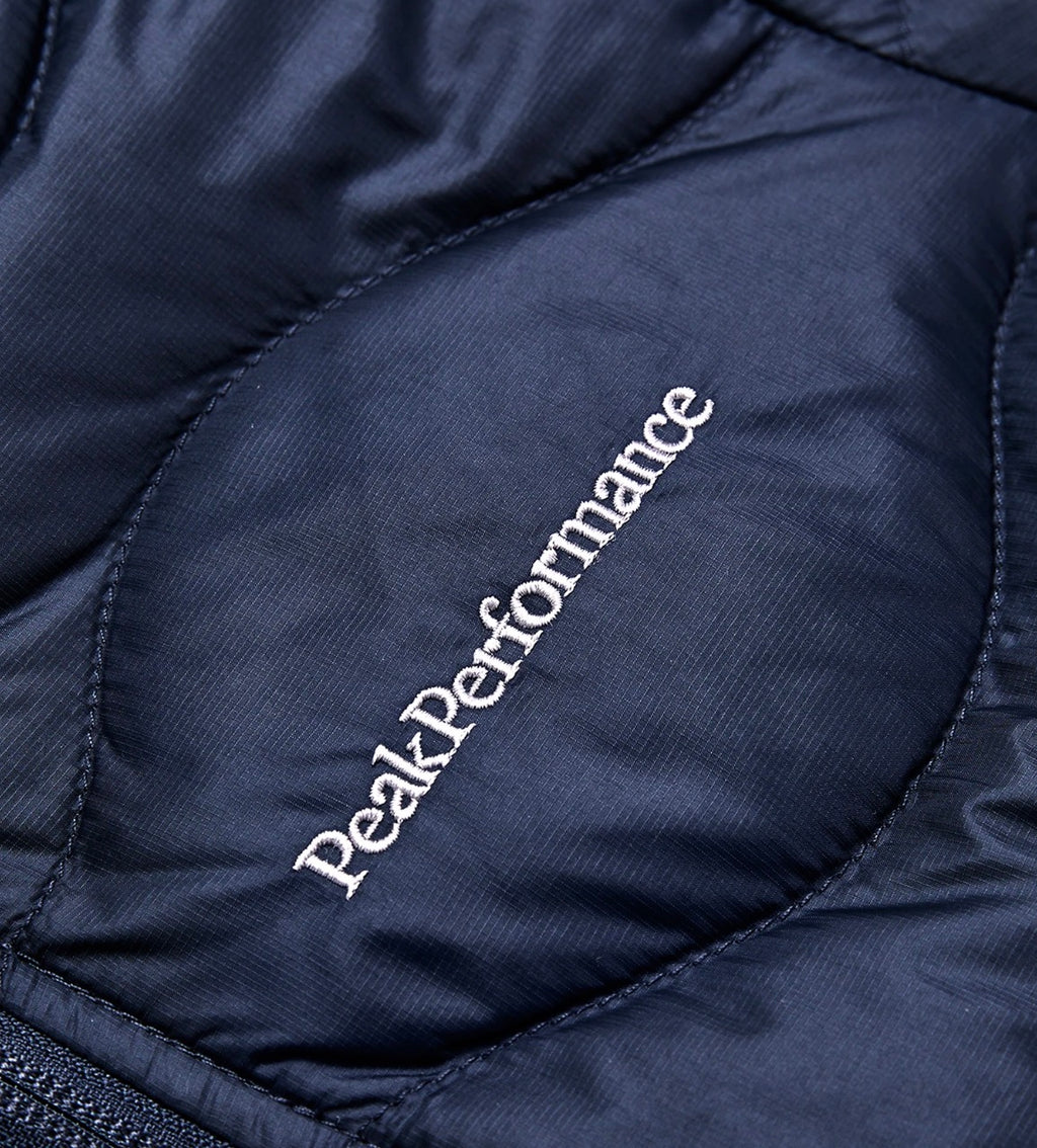Nærbilde av Peak Performance-logoen og quiltingen på den mørkeblå Jr Helium-dunjakken.