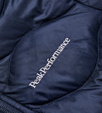 Nærbilde av Peak Performance-logoen og quiltingen på den mørkeblå Jr Helium-dunjakken.