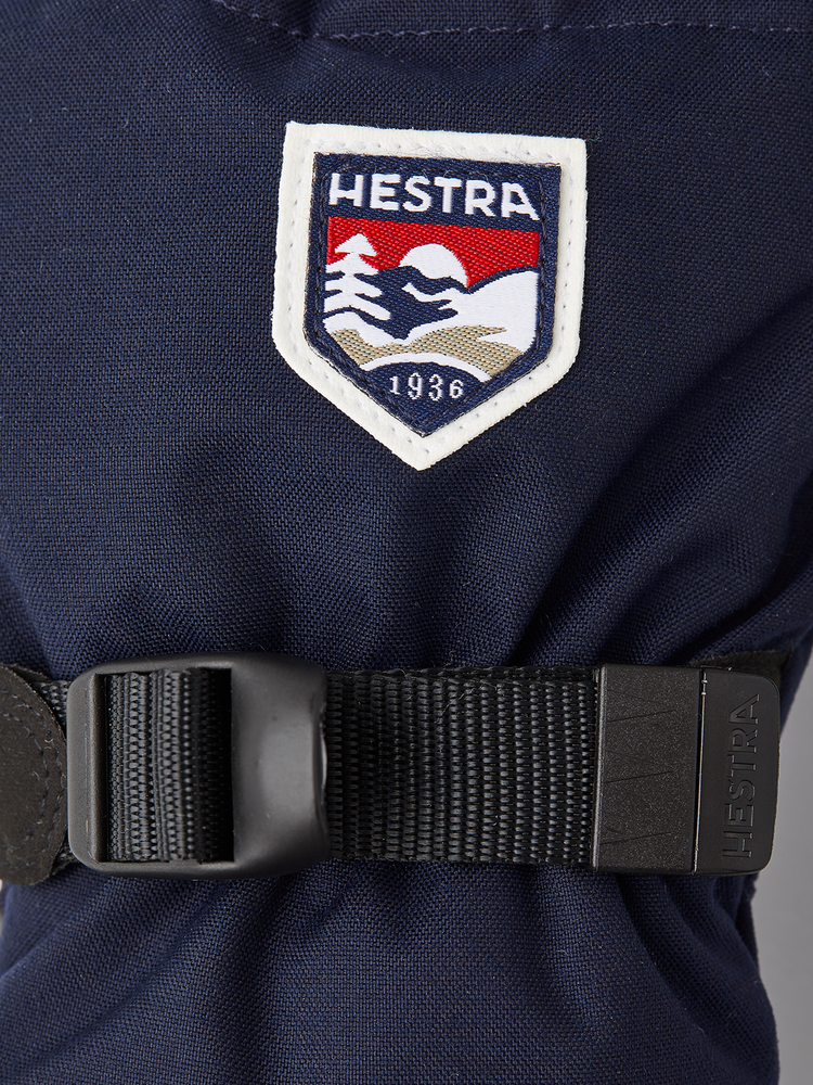 Nærbilde av logo og håndleddsrem på mørkeblå Hestra Fjellvotten Jr.